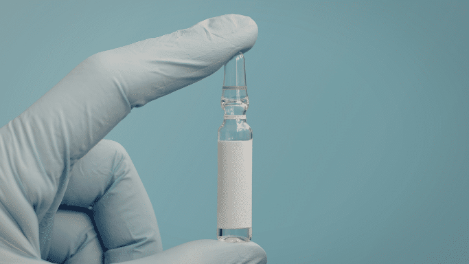 A gloved hand (light blue surgical glove) holding a small, clear glass ampoule with a blank white label between the thumb and forefinger.