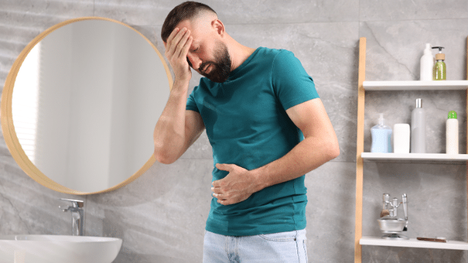 a man in a modern bathroom experiencing physical discomfort. He has a pained expression with his eyes closed, holding one hand to his forehead and the other over his stomach, suggesting he is suffering from a headache, nausea, or abdominal pain.