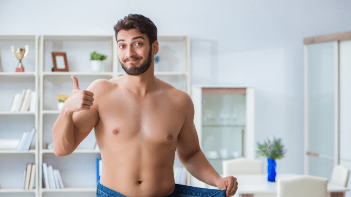 A shirtless man with a dark beard and hair stands indoors, smiling and giving a thumbs-up with one hand while holding out the waistband of his oversized jeans with the other, illustrating significant weight loss.