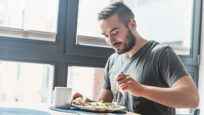 A young man with a short beard and styled hair sitting at a table by a window, focused on his meal. He is holding a fork and eating a healthy-looking breakfast consisting of eggs, greens, and a side of toast.