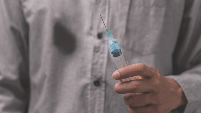 A close-up, slightly desaturated shot of a person’s hand holding a medical syringe filled with a clear blue liquid. The person is wearing a gray button-down shirt, and the focus is sharp on the needle.