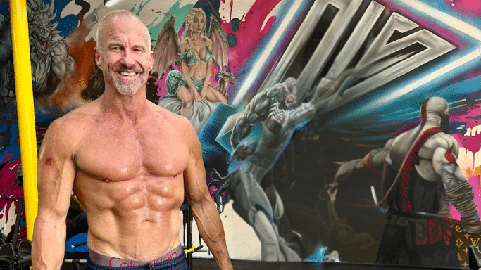A high-angle portrait of the same muscular gray-haired man from previous images, posing shirtless in a gym. He is showing off defined abdominal muscles and vascularity in his arms. The background features vibrant, comic-book-style mural art on the walls.