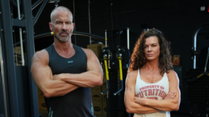 A medium shot of a muscular man with a shaved head and graying goatee standing next to a muscular woman with long, curly dark hair. Both have their arms crossed and are looking seriously at the camera inside a gym.