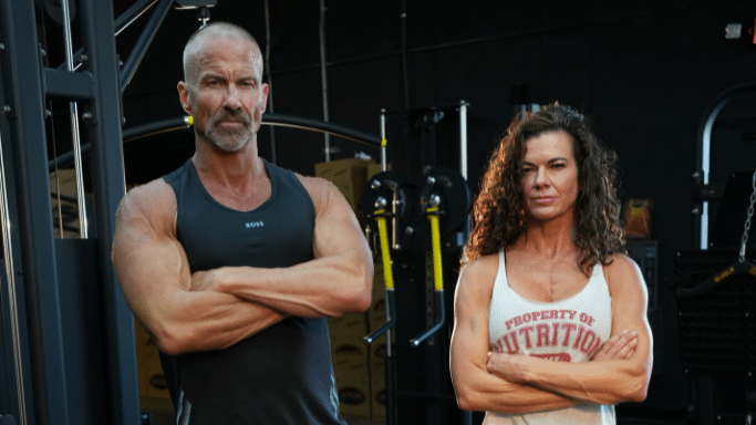 A medium shot of a muscular man with a shaved head and graying goatee standing next to a muscular woman with long, curly dark hair. Both have their arms crossed and are looking seriously at the camera inside a gym.