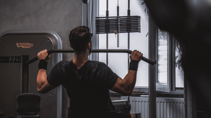 A view from behind of a man performing a lat pulldown on a cable machine. He is wearing a black t-shirt and wrist wraps, facing a window in a brightly lit gym.