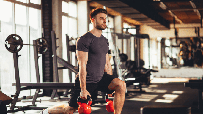 A man in a gray t-shirt and black shorts performing walking lunges while holding two orange kettlebells. The gym has large windows in the background letting in bright sunlight.