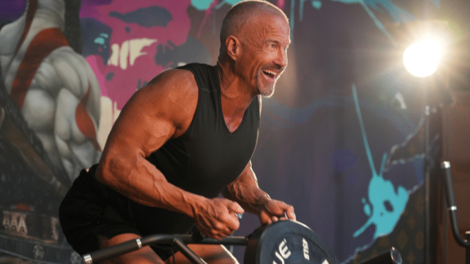 The muscular man smiling broadly while using a plate-loaded rowing machine in front of a colorful graffiti wall.