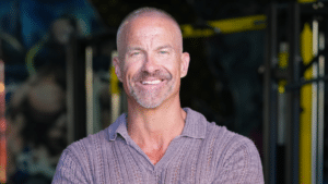 A medium-close-up portrait of a fit, middle-aged man with a shaved head and grey goatee, smiling warmly while wearing a textured purple polo shirt.