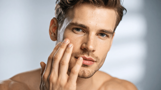 close-up portrait of a young man with styled brown hair, touching his cheek with his fingers as if applying skincare or checking his skin.