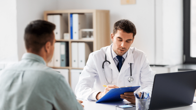 A wide shot of a doctor in a consultation room, sitting at a desk with a laptop and writing on a clipboard while talking to a patient seen from the back.