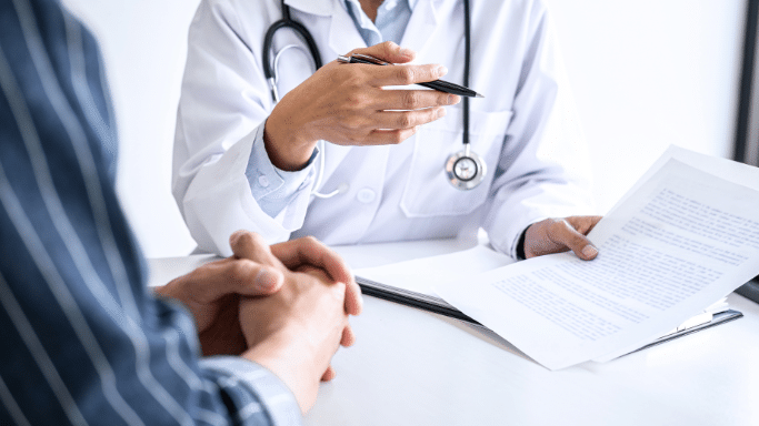 A close-up of a doctor in a white lab coat and stethoscope, holding a pen and pointing toward a document while consulting with a patient (only hands and torso of both are visible).
