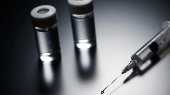 A close-up, dramatic shot of two glass vials with white caps and a medical syringe in the foreground. A single droplet of clear liquid hangs from the tip of the needle.