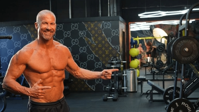 A fit, muscular middle-aged man with a shaved head and graying beard, smiling and pointing toward gym equipment. He is shirtless, and the gym wall behind him features a designer-patterned wallpaper.
