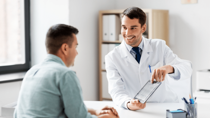 male doctor in a white lab coat sitting at a desk, smiling as he shows a digital tablet to a male patient. The patient is seen from the back, and the office is bright and professional.