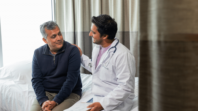 A male doctor in a white lab coat with a stethoscope around his neck sits on a hospital bed, smiling and placing a hand on the shoulder of a middle-aged male patient in a navy sweater. They are having a supportive conversation in a brightly lit room.