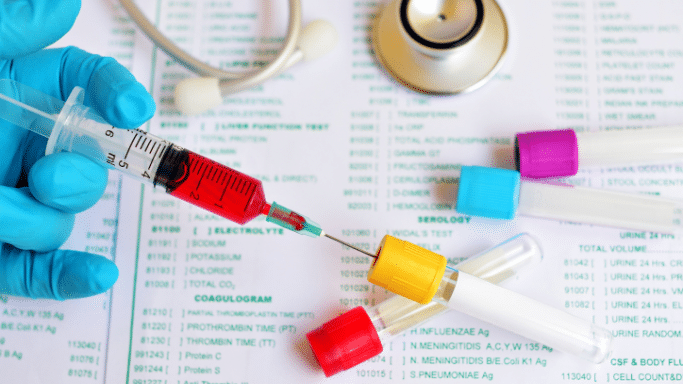 close-up, top-down view of a gloved hand holding a syringe filled with red liquid (blood) over a collection tube. The background shows a medical lab report with various test categories