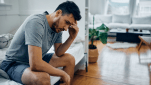 A man sits on the edge of a bed, his head resting in one hand with a discouraged and weary expression.