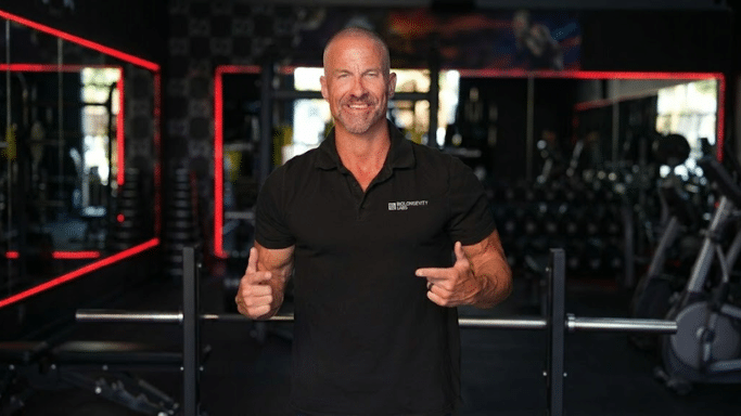 A muscular, bald man with a confident smile stands in a gym wearing a black polo shirt, gesturing toward himself with both thumbs.