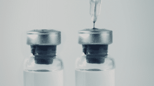 A close-up view of two glass medical vials with silver caps. A needle from a syringe is inserted into the top of the right vial.