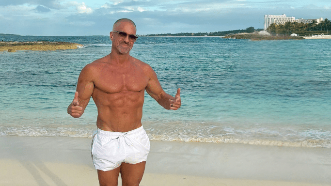 A shirtless, muscular man in white swim trunks and sunglasses stands on a tropical beach, smiling and giving two thumbs up.