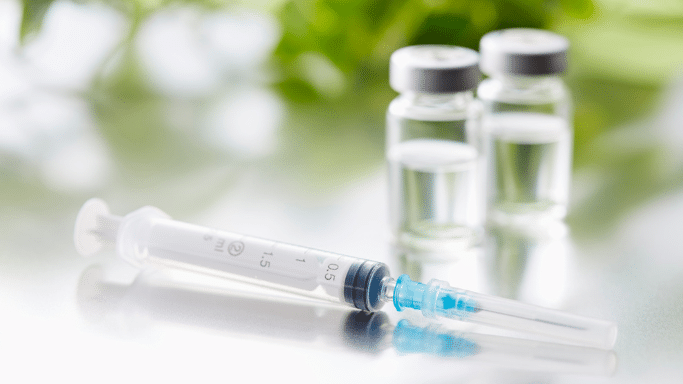 A clear medical syringe lies on a reflective surface in the foreground, with two small glass vials blurred in the background against a soft green backdrop.