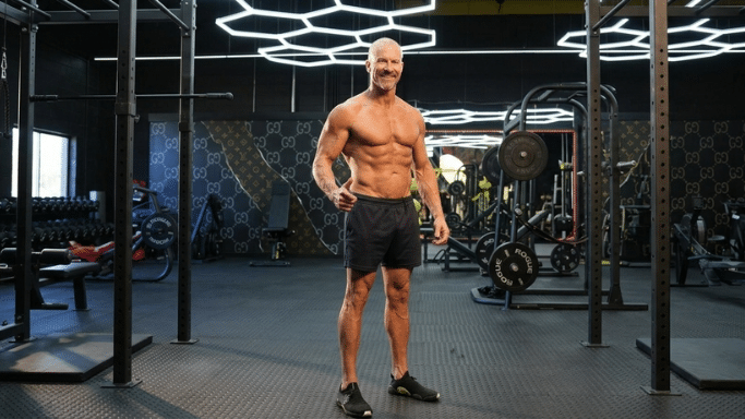 A full-body shot of a muscular, shirtless man standing in a modern, high-end gym. He is smiling and showcasing a lean, athletic physique.