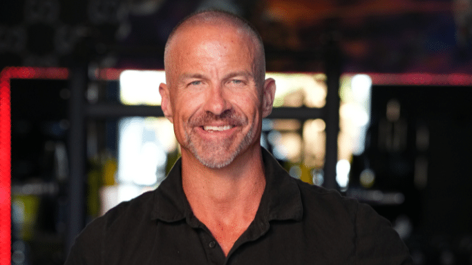 A head-and-shoulders portrait of a smiling, middle-aged man with a short beard and a bald head, wearing a black polo shirt in a gym setting.
