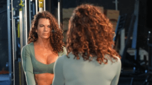 A medium shot of a woman with long, curly brown hair looking at her reflection in a gym mirror. She is wearing a sage green long-sleeved crop top.