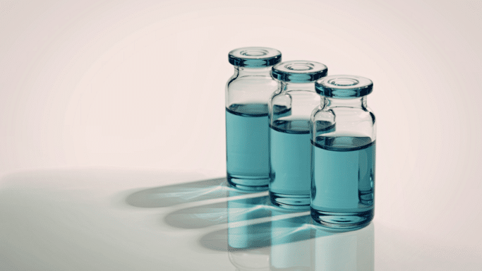 Three small glass medical vials filled with a clear blue liquid, arranged in a diagonal line on a white surface. Sharp shadows are cast to the left of each vial.