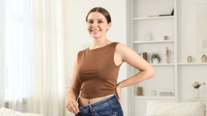 A smiling woman in a brown sleeveless top standing in a living room, pulling the waistband of her blue jeans away from her waist to show significant weight loss.