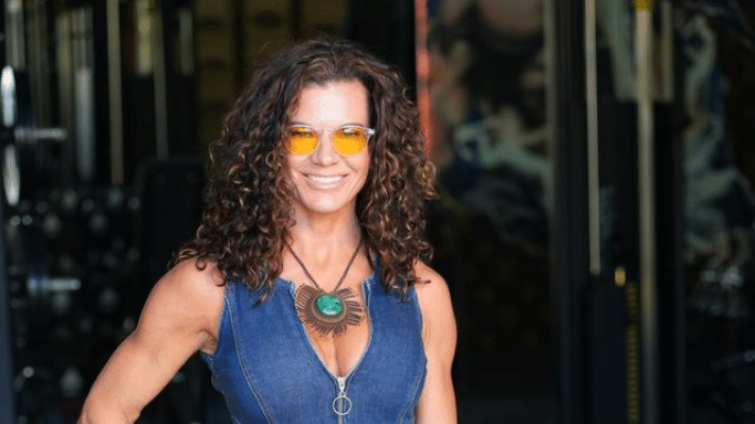 A portrait of the same athletic woman from previous images, smiling and wearing yellow-tinted glasses and a denim vest. Her curly hair is well-defined, and she is standing in a gym environment.