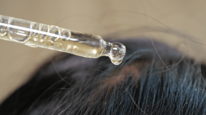 A macro shot showing a glass dropper releasing a single clear, golden drop of oil or serum onto a person's scalp and dark hair.