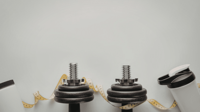 A minimalist fitness-themed flat lay against a light grey background. In the lower half, there are two black adjustable dumbbells, a yellow measuring tape curled around them, and two white protein shakers on either side. The top half of the image is empty space.