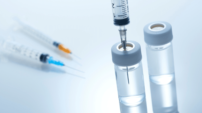 A close-up, high-angle shot of a medical setting. A syringe is shown with its needle inserted through the rubber stopper of a clear glass vial containing liquid. A second vial and two more syringes with orange and blue caps lie blurred in the background on a clean, reflective white surface.