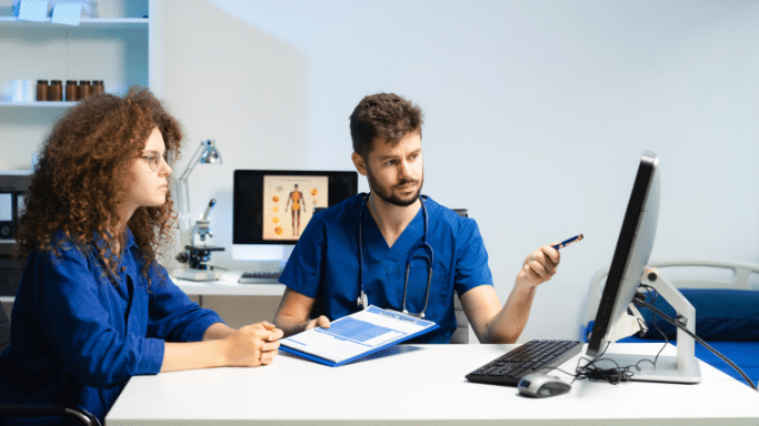 A male doctor in blue scrubs sits at a white desk in a clinical setting, pointing a pen toward a computer monitor. He is speaking to a woman with curly hair who sits across from him.