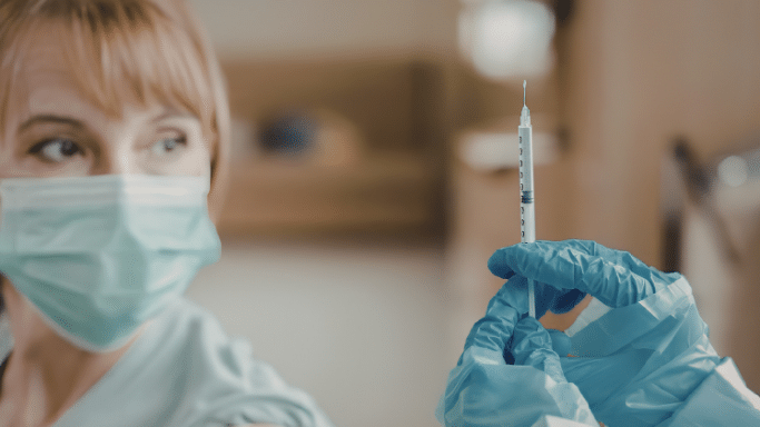 A close-up of a healthcare professional wearing a light green surgical mask and blue medical gloves. They are holding up a small syringe, carefully inspecting the needle and the fluid inside.