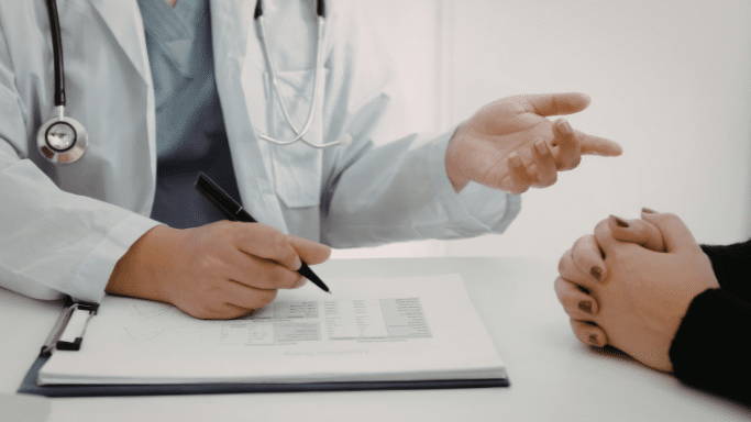 A mid-section view of a consultation. A doctor in a white lab coat and blue scrubs holds a black pen over a medical document on a clipboard. They are gesturing with their other hand while talking to a patient whose hands are clasped on the table.