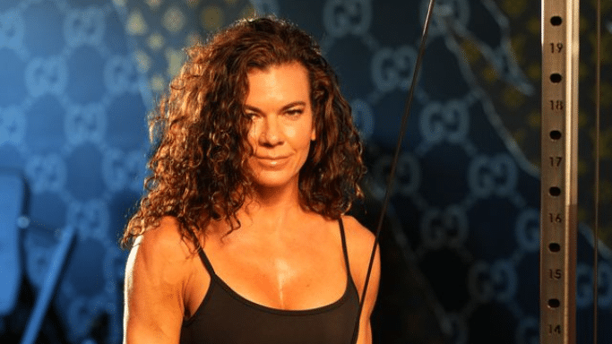 A portrait of a woman with curly brown hair in a gym setting. She is wearing a black tank top and looking directly at the camera with a subtle smile. A gym cable machine is visible to the right, and the background features a wall with designer-style logos.