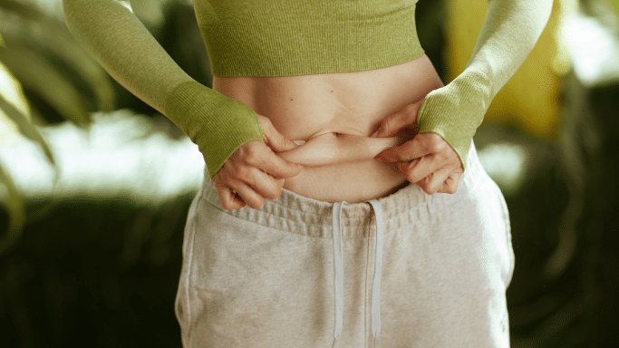 A mid-section view of a person wearing a lime green long-sleeved athletic top and grey sweatpants. They are using their fingers to pinch a small amount of skin/fat on their abdomen, highlighting body composition.