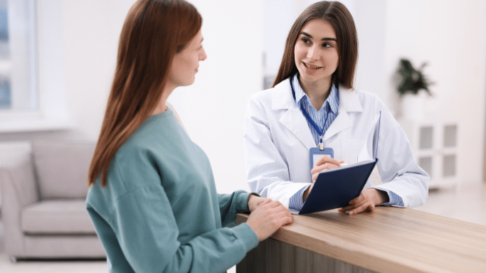 A professional medical setting where a young female healthcare worker in a white lab coat and blue striped shirt is smiling and speaking with a female patient. The clinician is holding a blue folder and a pen while leaning against a wooden counter.