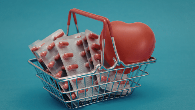 A miniature wire shopping basket with red handles, placed against a solid teal background. Inside the basket are several silver blister packs containing red capsules, leaning against a smooth, red heart-shaped object.