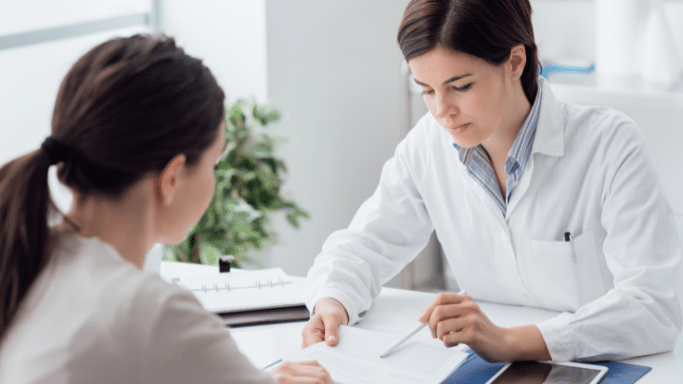 A female doctor in a white lab coat sits at a desk, pointing with a pen to a document she is reviewing with a patient. The patient is seen from the back, showing only their dark hair tied in a ponytail. The setting is a bright, professional medical office.