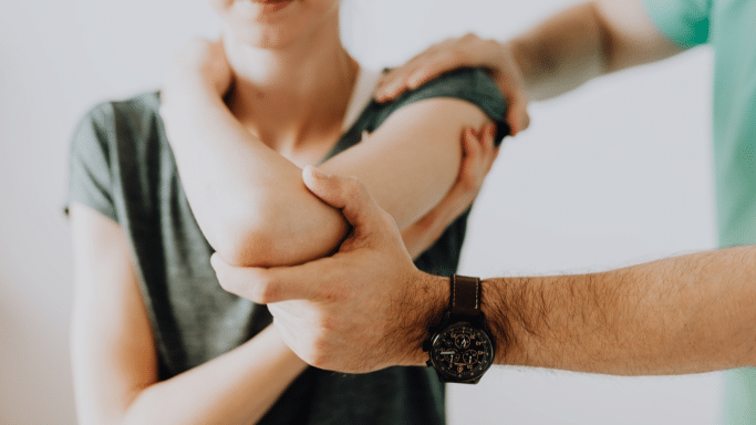 A close-up shot of a physical therapy or medical examination. A person in a green shirt is using their hands to stabilize and assess a patient's elbow and arm. The practitioner is wearing a large brown leather watch.