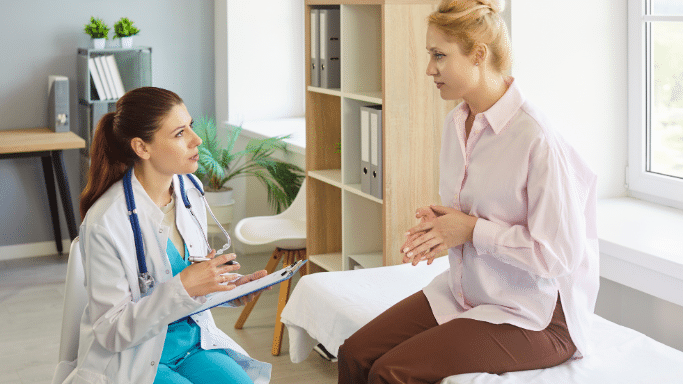 A female doctor with her hair in a ponytail, wearing a white coat over blue scrubs, sits with a clipboard and consults with a blonde female patient. The patient is sitting on an examination table, looking concerned while gesturing with her hands.