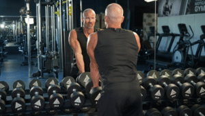 A muscular, bald man in a black sleeveless shirt viewed from behind, looking at his reflection in a gym mirror. In front of him is a long rack filled with various dumbbells labeled with their weights.