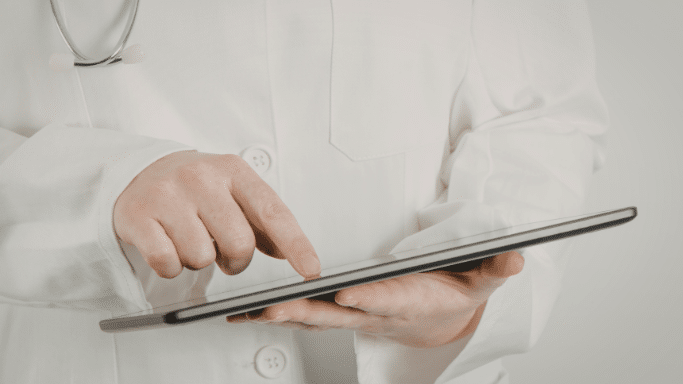 A close-up of a person wearing a white medical lab coat. A stethoscope hangs around their neck, and they are using an index finger to navigate a thin, black tablet held in their hands.