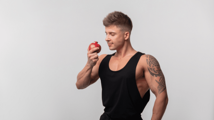 A muscular man with a tribal tattoo on his shoulder and arm stands against a plain light grey background. He is wearing a black tank top and is holding a red pomegranate up to his face, looking at it with a slight smile.