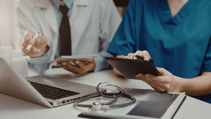 A high-angle, close-up shot of two medical professionals working at a desk. One is in a white coat and tie, pointing with a pen, while the other is in teal scrubs using a tablet. A laptop, stethoscope, and medical folders are spread across the workspace.