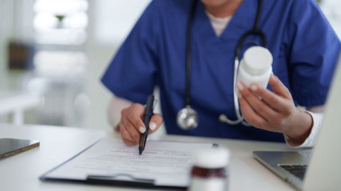 A close-up, slightly blurred shot of a healthcare professional in blue scrubs sitting at a desk. One hand holds a white medicine bottle while the other uses a pen to point at a document on a clipboard. A laptop and stethoscope are also visible.