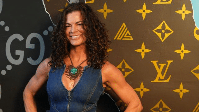 A portrait of a smiling woman with voluminous, curly brown hair. She is wearing a denim vest and a large green stone necklace. She stands in front of a dark backdrop decorated with gold-colored logos, including the Louis Vuitton monogram.